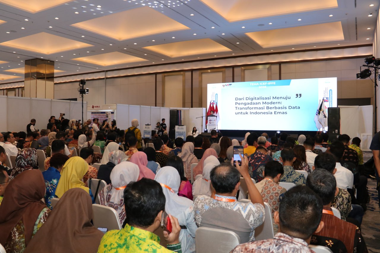 Peserta duduk menghadap panggung dan layar presentasi dalam sebuah forum.
