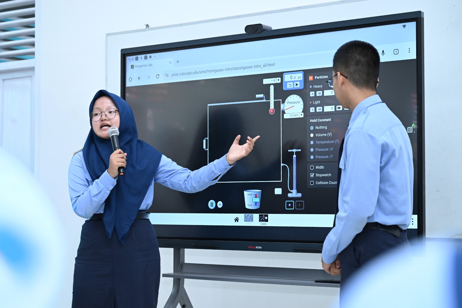 Dua siswa mempresentasikan simulasi sains pada papan interaktif digital di ruang kelas.